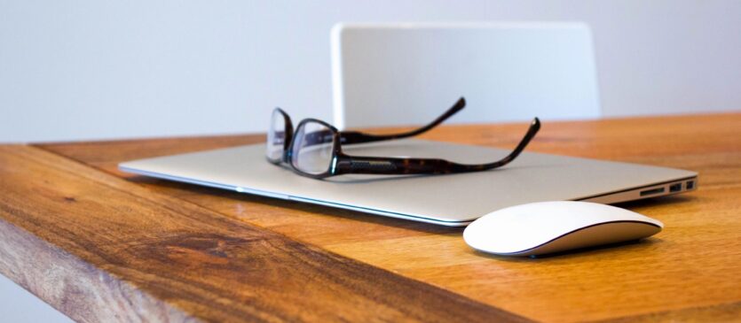 A laptop on a wooden desk with glasses and a mouse.